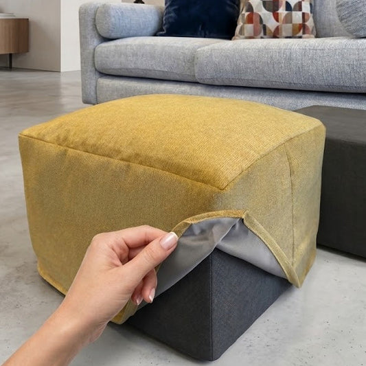 Yellow ottoman with a hand holding a clear adhesive strip in a living room setting.