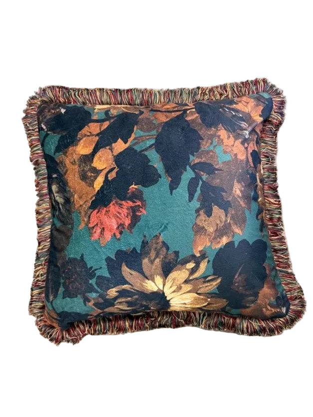 A square decorative throw pillow with a multicolor floral pattern on a teal background and fringed edges, placed on a beige carpet.