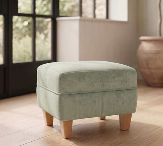 Green ottoman with wooden legs on a wooden floor in a room with large windows.